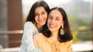 Two women smiling together indoors near a window in a bright, modern setting.