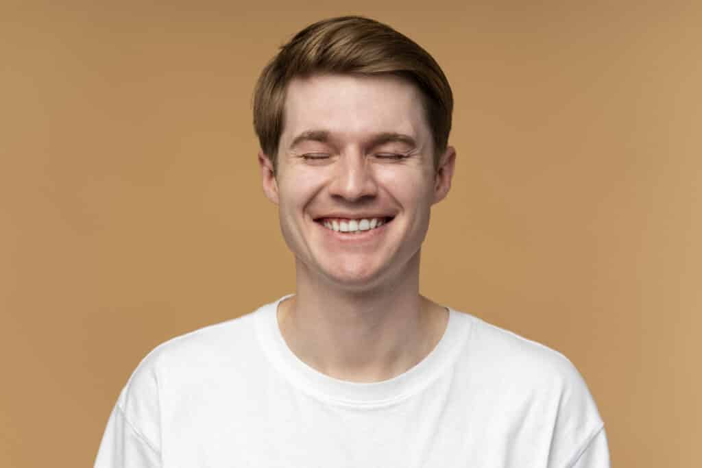 Young man smiling at camera wearing white shirt against tan background, professional headshot.