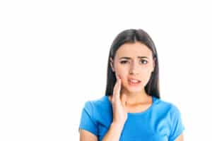Young woman in blue shirt touching face, appearing thoughtful about dental concerns against white background.