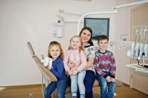 Dentist holding three smiling children in dental office treatment room with equipment visible.