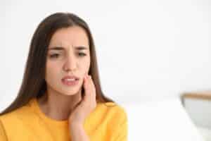 Woman in yellow shirt touching cheek, appearing to experience tooth pain or discomfort.