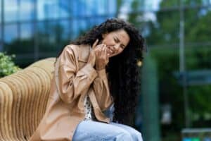Woman with hand to face experiencing tooth pain, seated outdoors near modern building.